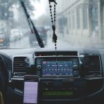 Motorista ao volante usa o celular para navegação enquanto percorre uma cidade sob chuva.
