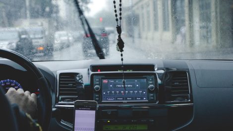 Motorista ao volante usa o celular para navegação enquanto percorre uma cidade sob chuva.