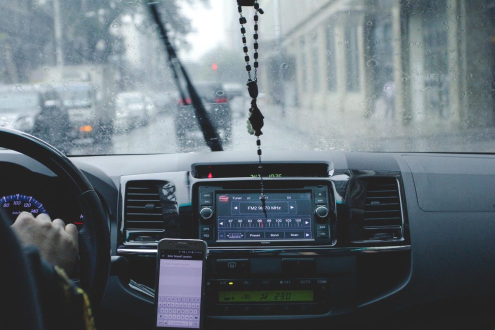 Motorista ao volante usa o celular para navegação enquanto percorre uma cidade sob chuva.