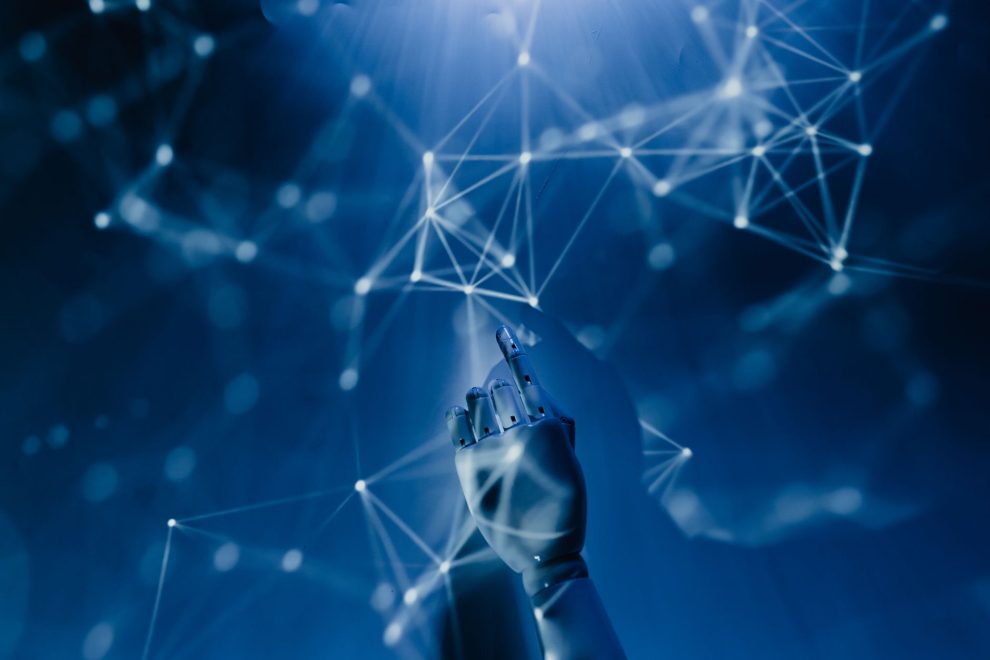 Mão robótica alcança uma rede digital azul, em representação conceitual da inteligência artificial e das tecnologias conectadas.