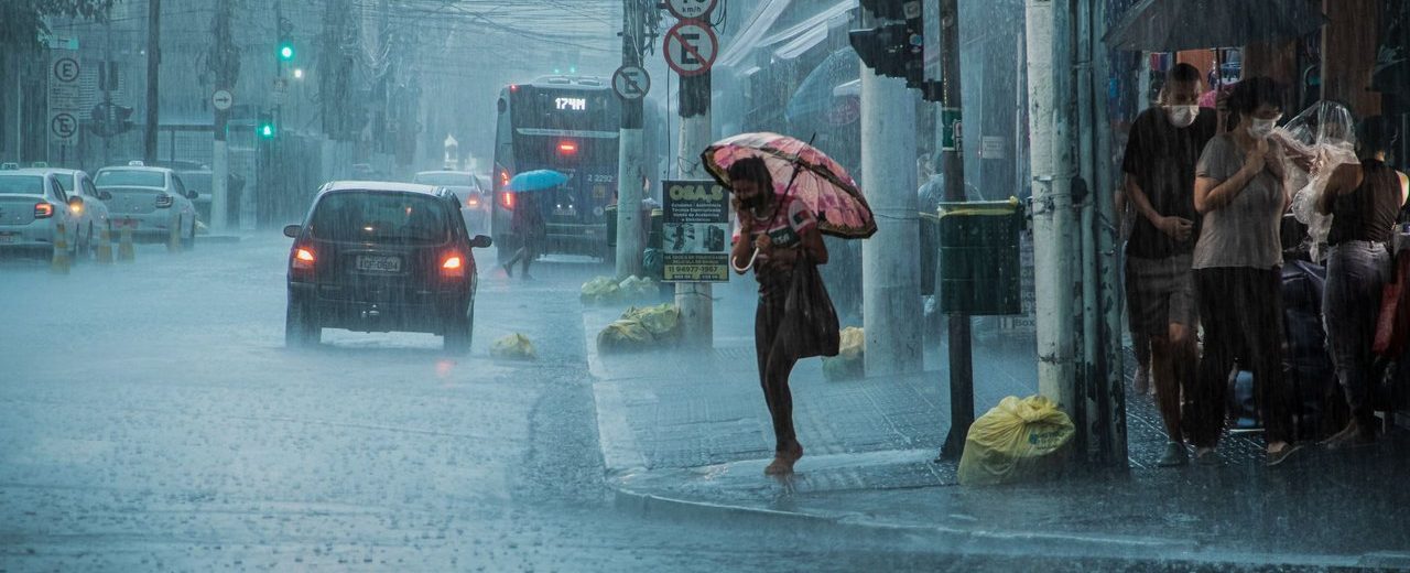 Rua movimentada durante chuva intensa, com carros, pedestres e pessoas usando guarda-chuvas.