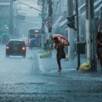 Rua movimentada durante chuva intensa, com carros, pedestres e pessoas usando guarda-chuvas.