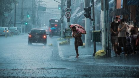 Rua movimentada durante chuva intensa, com carros, pedestres e pessoas usando guarda-chuvas.