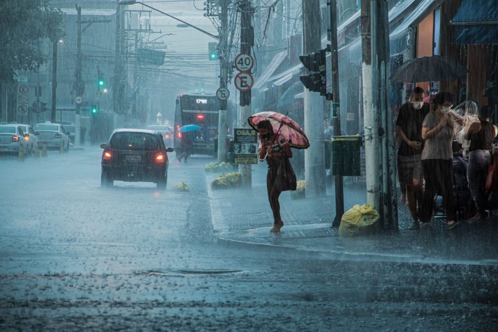 Rua movimentada durante chuva intensa, com carros, pedestres e pessoas usando guarda-chuvas.