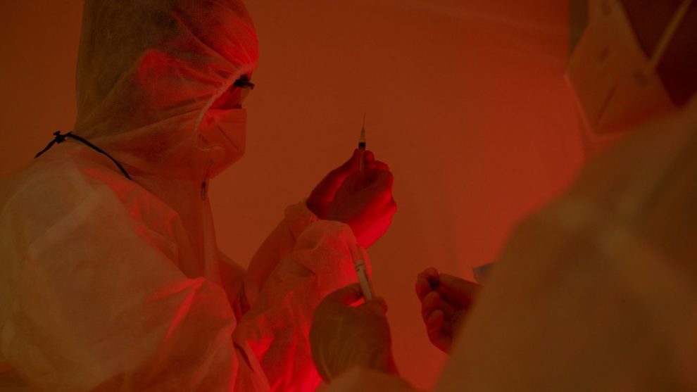 Pesquisadores usando roupas de proteção preparam seringas em um laboratório.