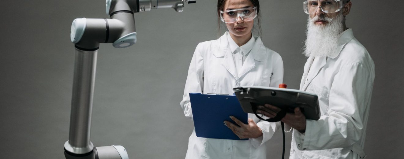 Dois pesquisadores de jaleco analisam um braço robótico em um laboratório.