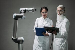 Dois pesquisadores de jaleco analisam um braço robótico em um laboratório.