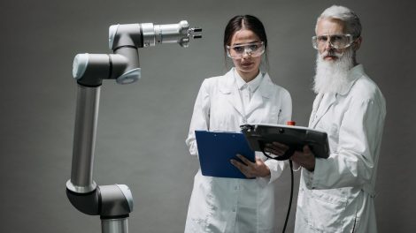 Dois pesquisadores de jaleco analisam um braço robótico em um laboratório.